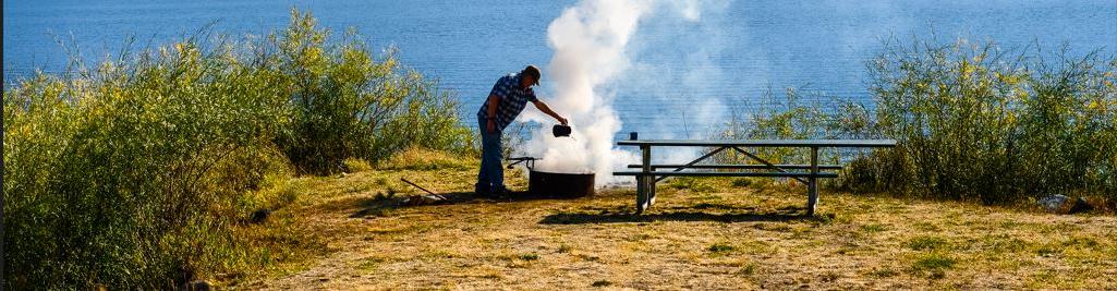 一名男子从一个茶壶里倒水，在一个篝火上，在一个湖前建立了一个篝火环，背景是山脉。